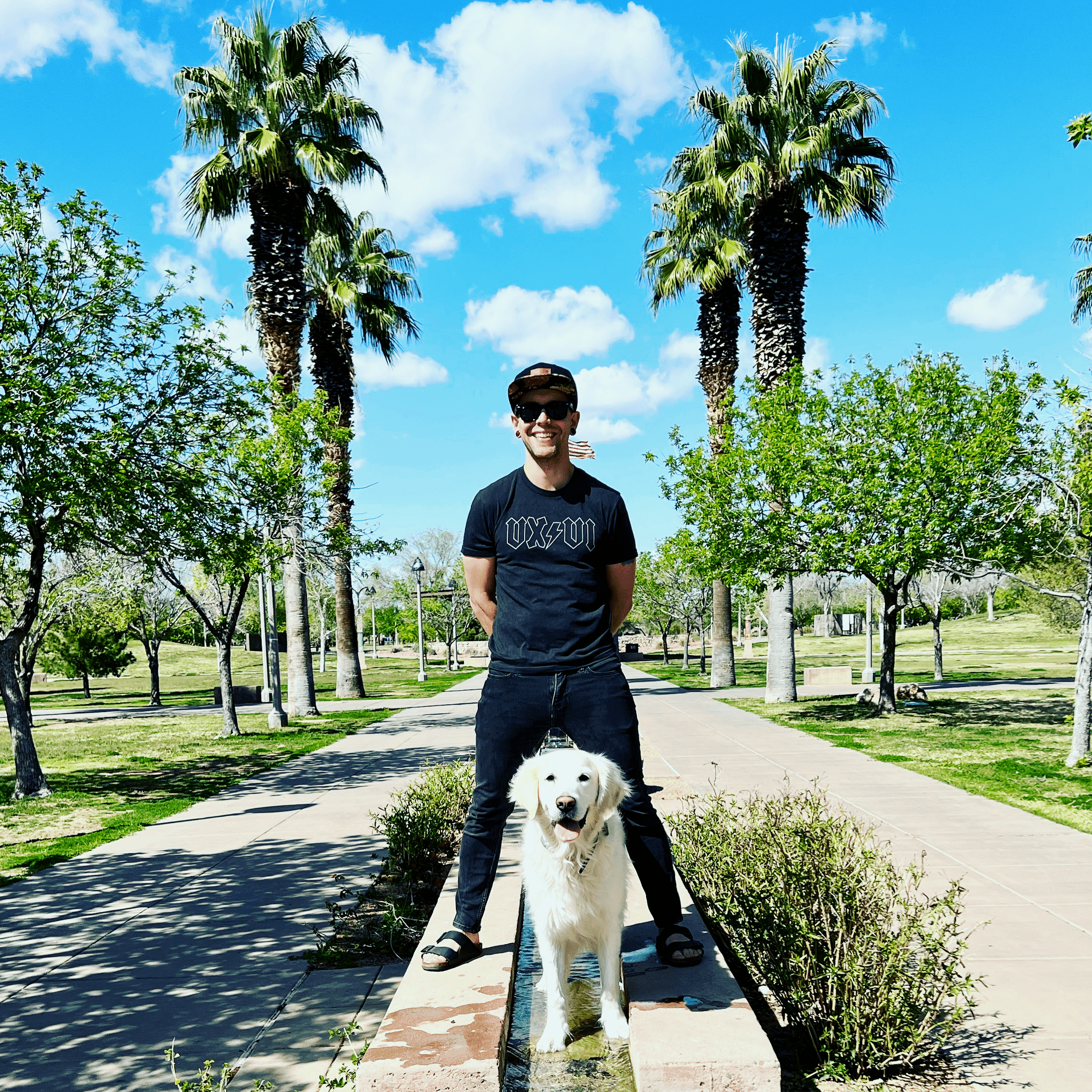 Zac Elletson standing on a path between two palm trees with a white golden retriever at his feet