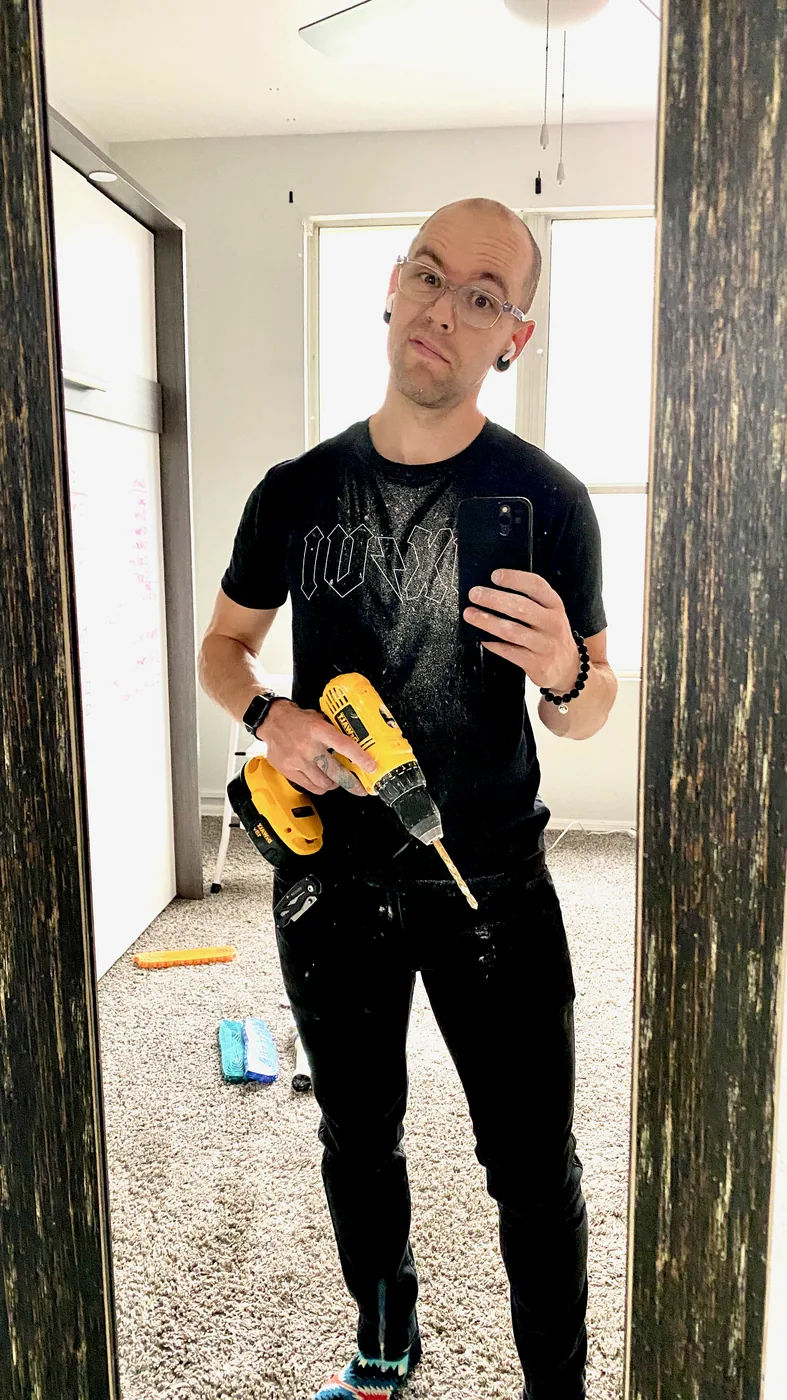 Mirror selfie of Zac in a paint-spattered t-shirt holding a DeWalt drill, standing in an unfinished room with bare carpet and walls mid-renovation