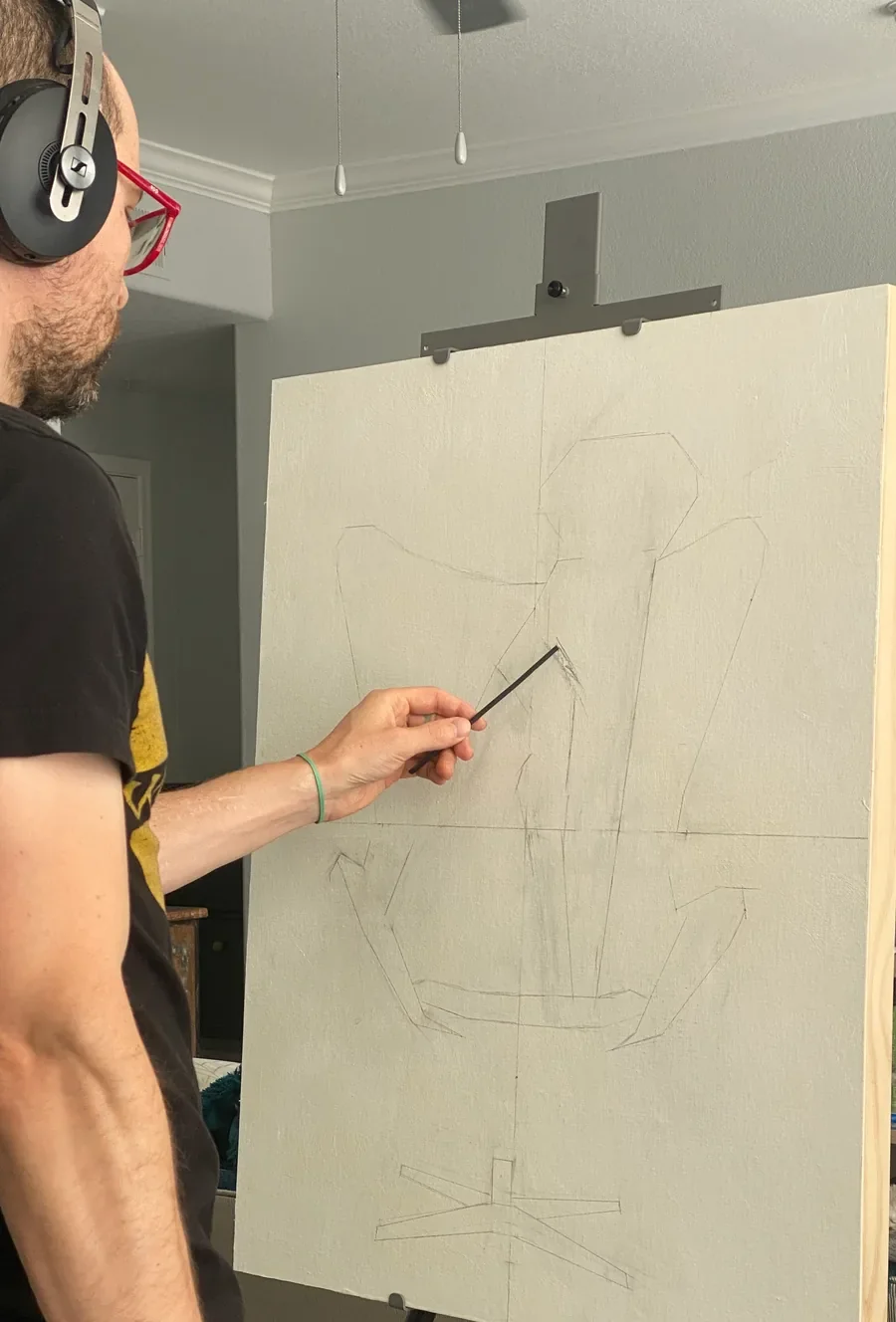 Zac at the easel with headphones, drawing an initial pencil sketch on a fresh canvas before paint