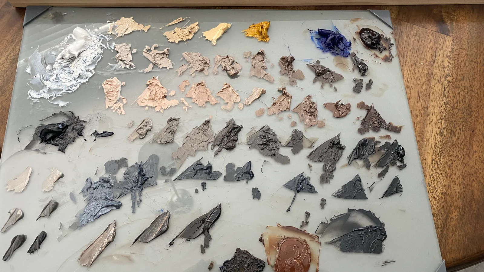 A glass palette with arrays of pre-mixed oil paint — flesh tones across the top, neutral greys through the middle, blues and umbers along the bottom
